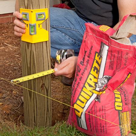 A square wooden post is inserted in the soil, a man is using the post level to plumb it.